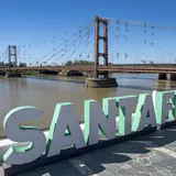 Letras corpóreas que forman la palabra SANTA FE en la costanera, con el histórico Puente Colgante y la laguna Setúbal de fondo bajo un cielo azul.